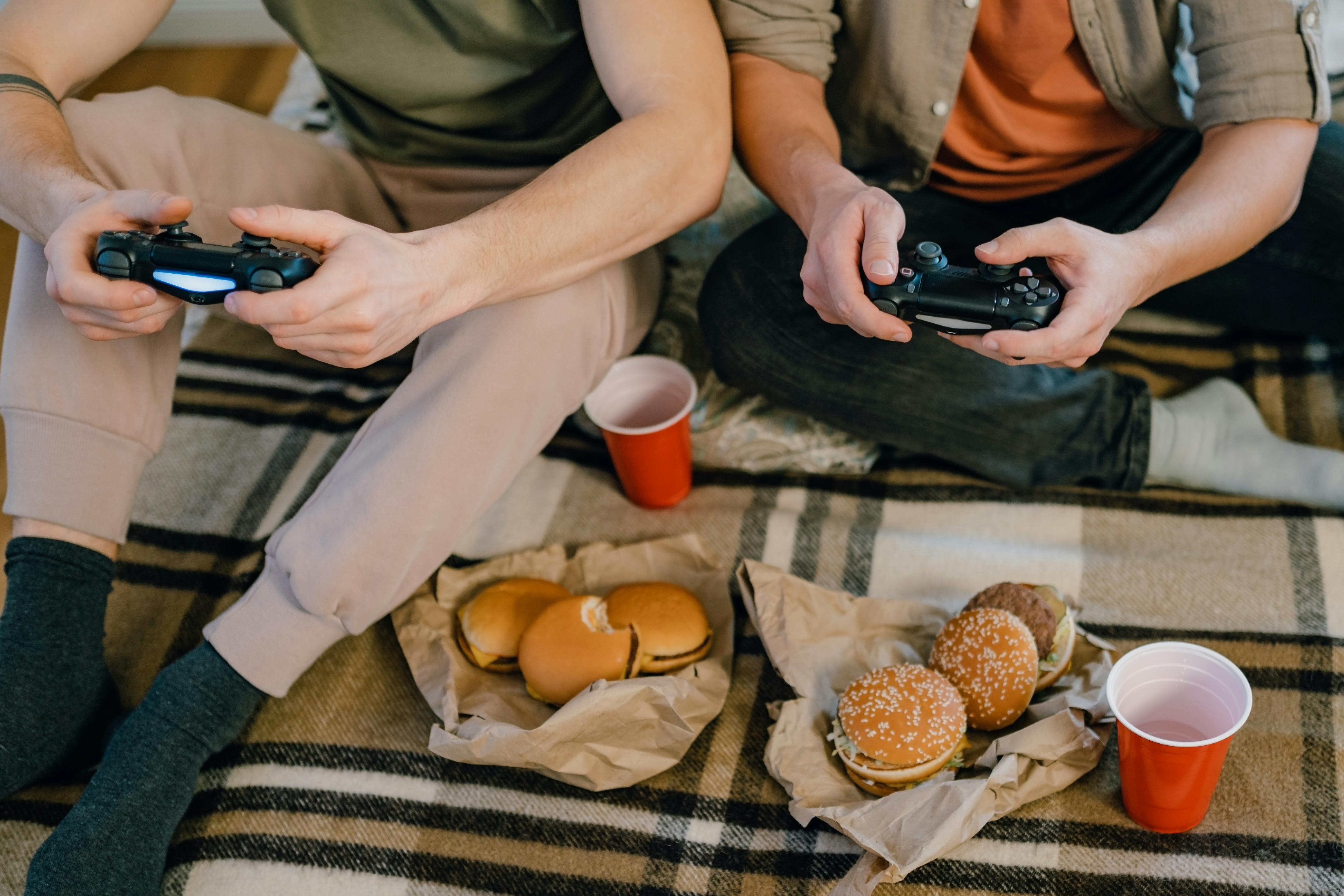 Dos hombres sentados en el suelo comiendo hamburguesas mientras juegan a videojuegos | Fuente: Pexels