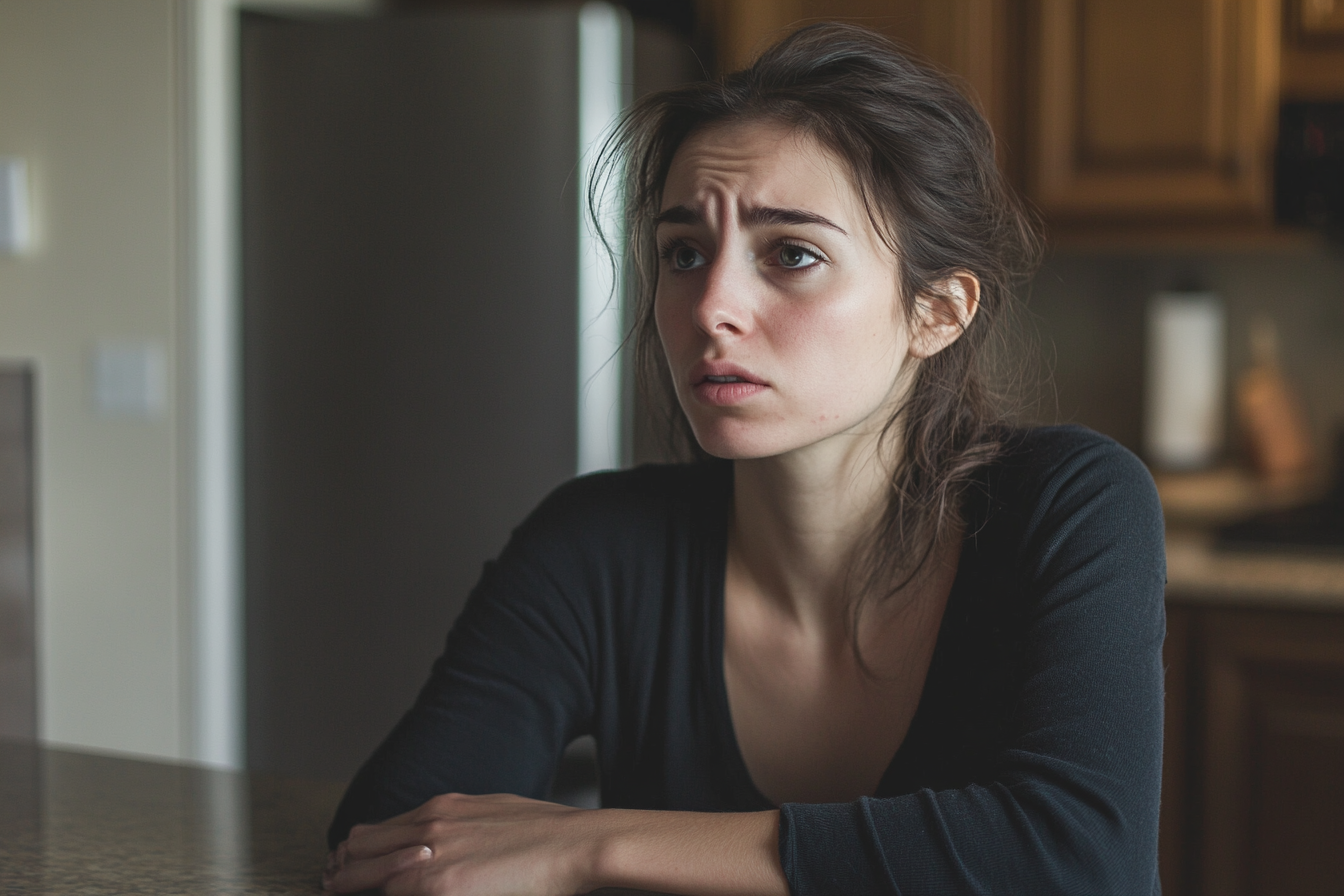 Una mujer triste y conmocionada sentada a la mesa de la cocina | Fuente: Midjourney