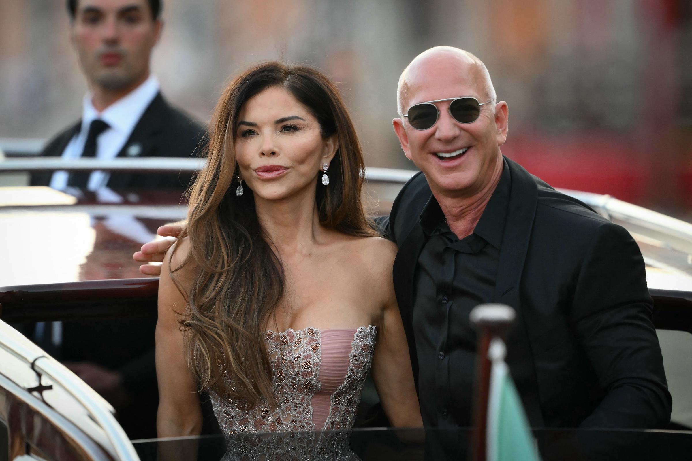 Jeff Bezos esboza una amplia sonrisa mientras se inclina hacia Lauren Sánchez, que mira al frente con expresión serena. La pareja está de pie en la cubierta del barco, rebosante de confianza, mientras un guardia de seguridad trajeado les observa desde atrás frente a un paisaje urbano borroso.