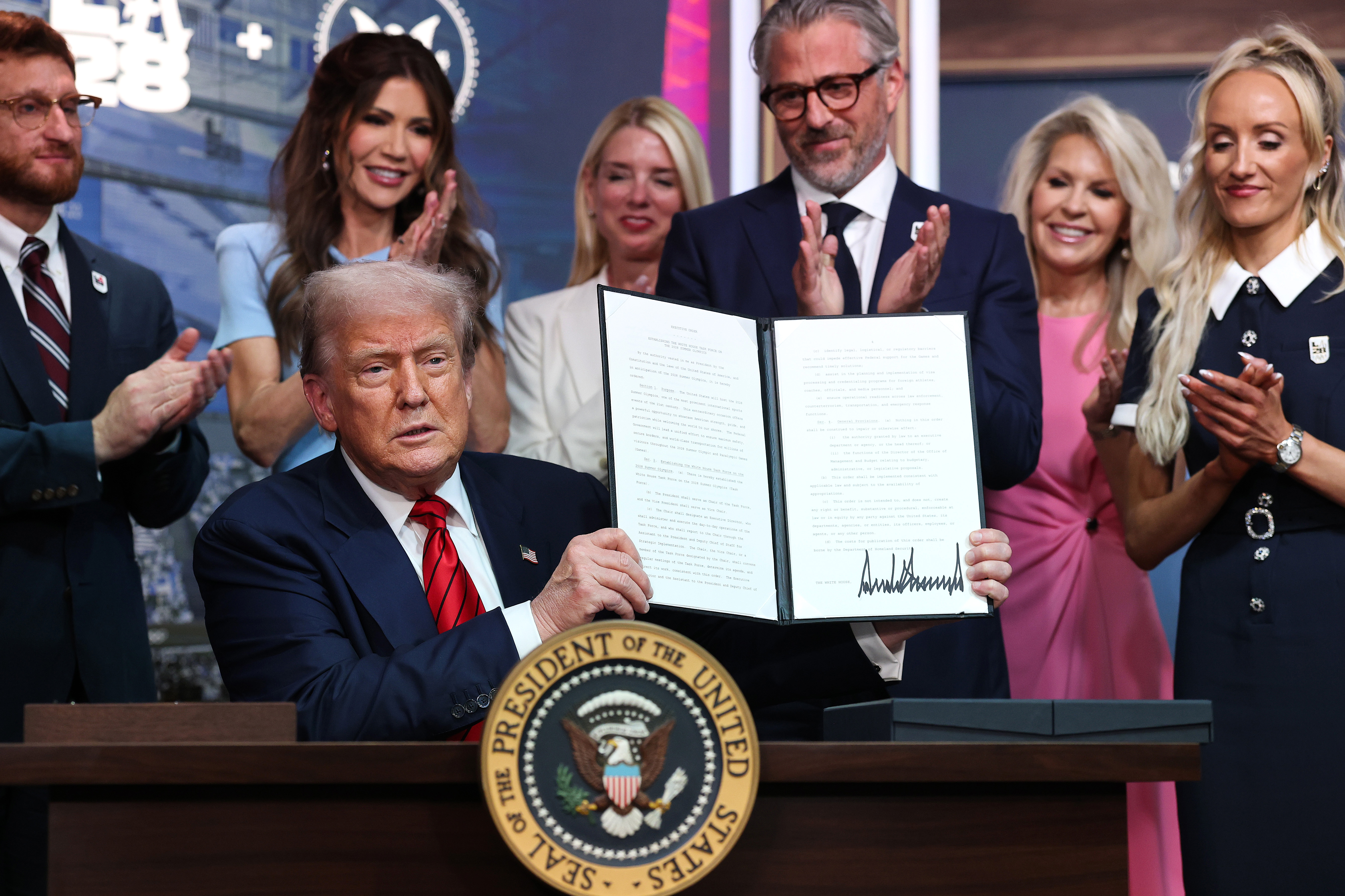Con la orden ejecutiva firmada en alto, Trump la presenta al público en el Auditorio South Court de la Casa Blanca, mientras funcionarios e invitados aplauden.