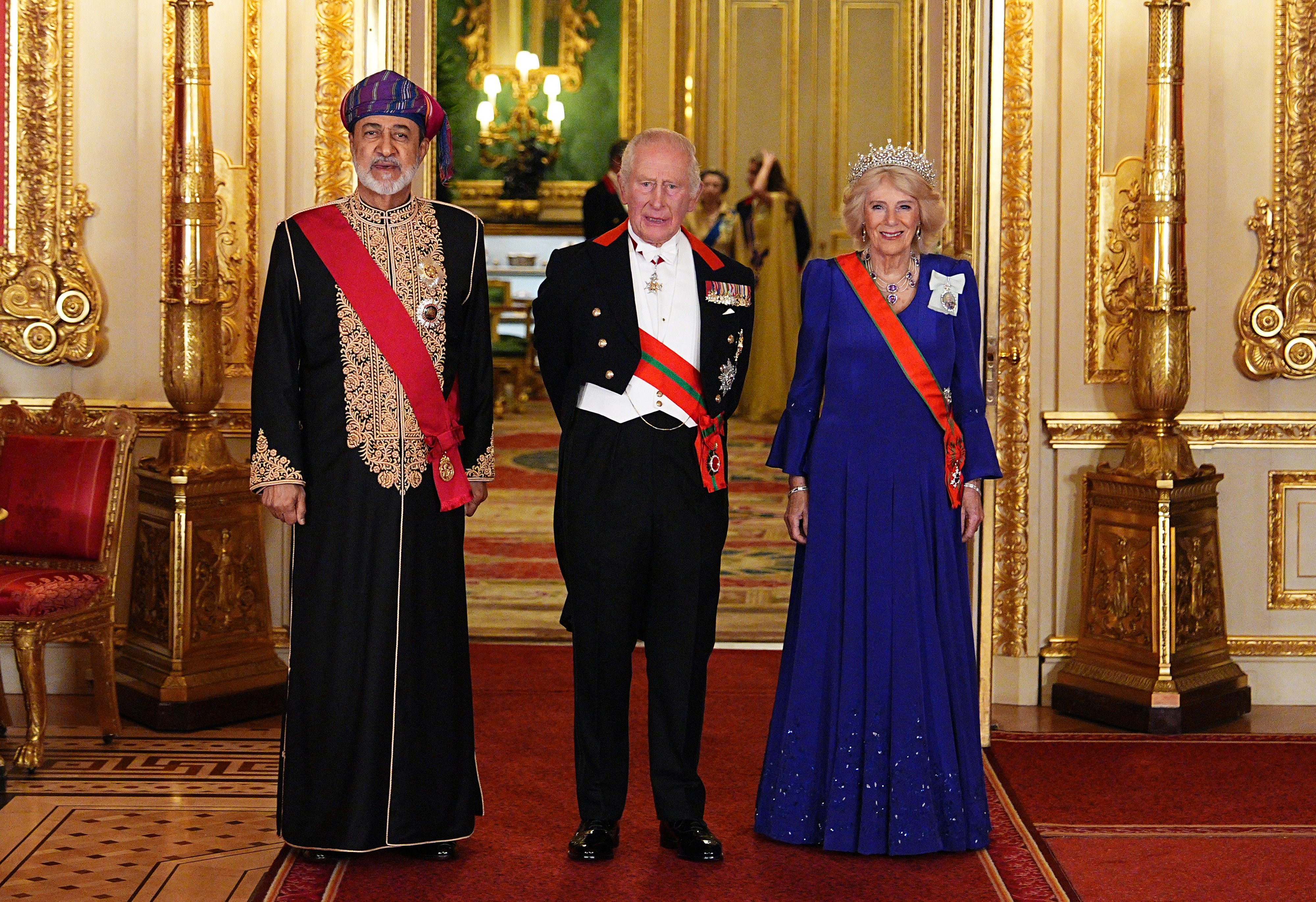 El sultán Haitham bin Tarik de Omán posa junto al rey Charles III y la reina Camilla en el castillo de Windsor, Inglaterra, antes del lujoso banquete de Estado del 20 de octubre de 2026. Rodeado de una ornamentada decoración dorada, Charles III viste frac con medallas militares y bandas de órdenes honoríficas. El sultán luce una túnica negra con elaborados bordados dorados, banda ceremonial roja y un colorido turbante tradicional massar. Camilla deslumbra con un vestido azul plisado, la brillante tiara de zafiros belgas y una banda naranja. | Fuente: Getty Images
