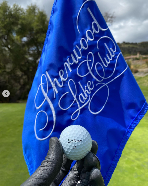 Brandon Thomas Lee sostiene una pelota de golf delante de una bandera azul impresa con el logotipo del Sherwood Country Club, publicado en marzo de 2021 | Fuente: Instagram/brandonthomaslee