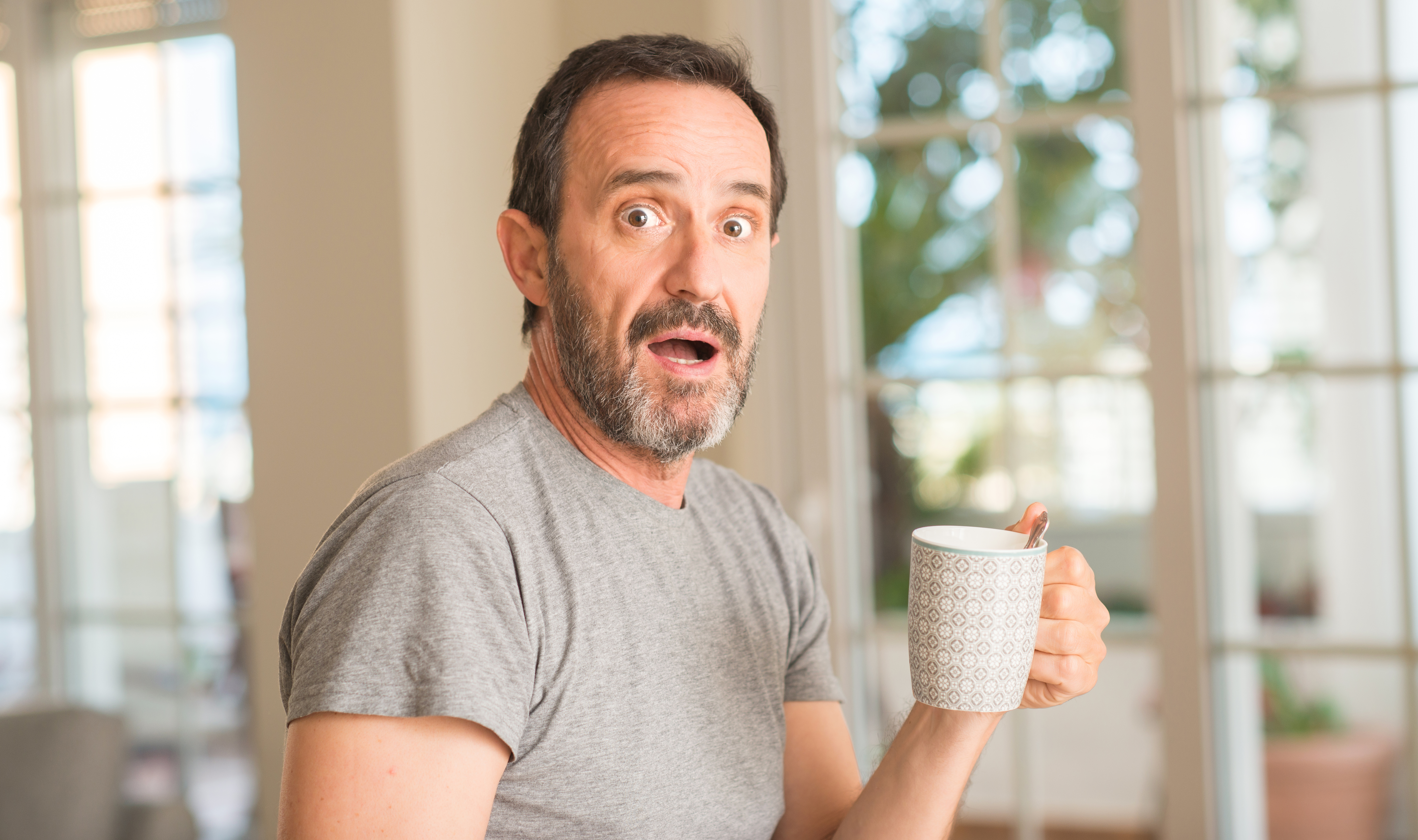 Hombre de mediana edad bebiendo café en una taza asustado con cara de sorpresa, asustado y excitado con expresión de miedo | Fuente: Getty Images