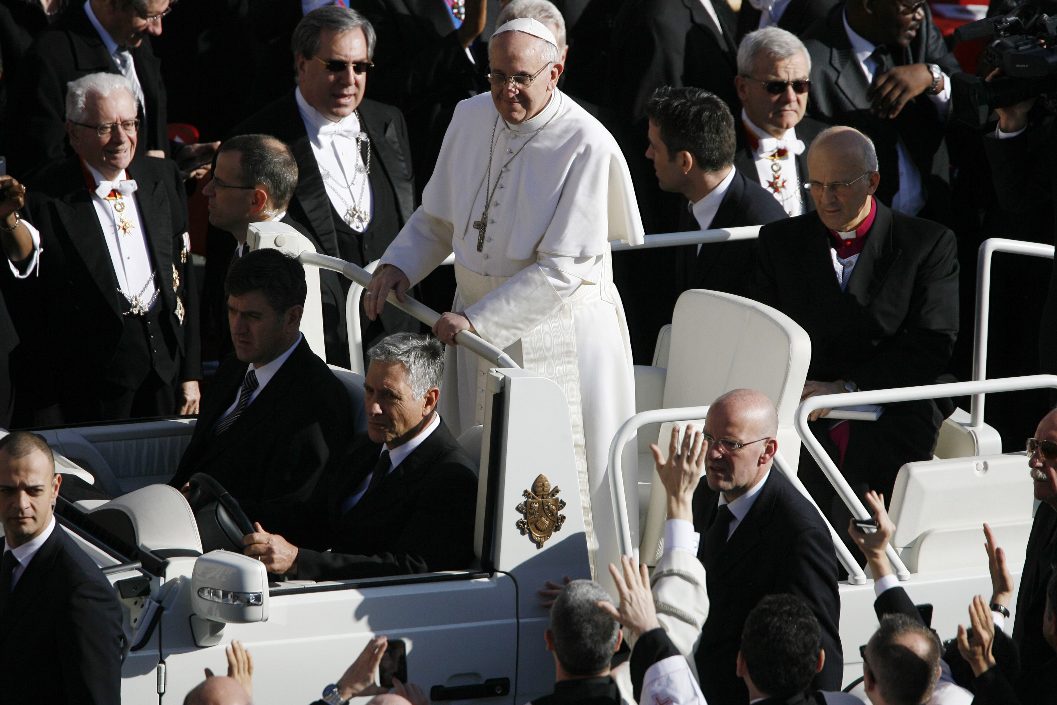 El papa Francisco, de pie en el papamóvil, rodeado de un mar de dignatarios y agitando las manos, vestido de blanco y con una cruz de plata sobre el pecho.