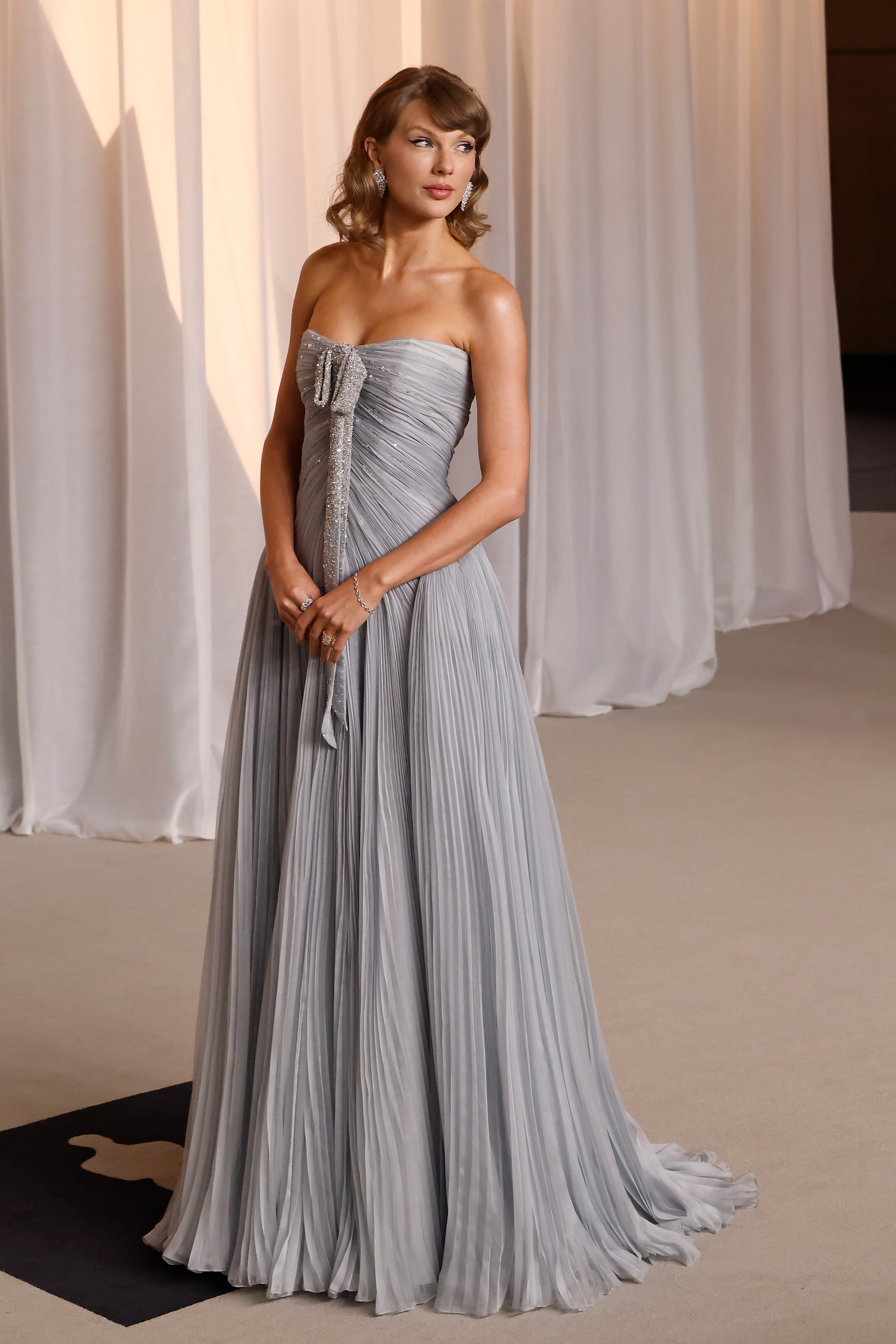 Taylor Swift irradia una elegancia etérea con un vestido sin tirantes de color gris paloma, realzado por delicados pliegues en todo el corpiño y la falda con gran vuelo. Un brillante lazo adornado con cristales cae con elegancia por la parte delantera, complementado con unos pendientes de diamantes en forma de gota y un suave peinado con ondas de estilo vintage. Lista para subir al escenario como la artista musical destacada de la noche, la ganadora de un Grammy desprende un glamour puro y atemporal.