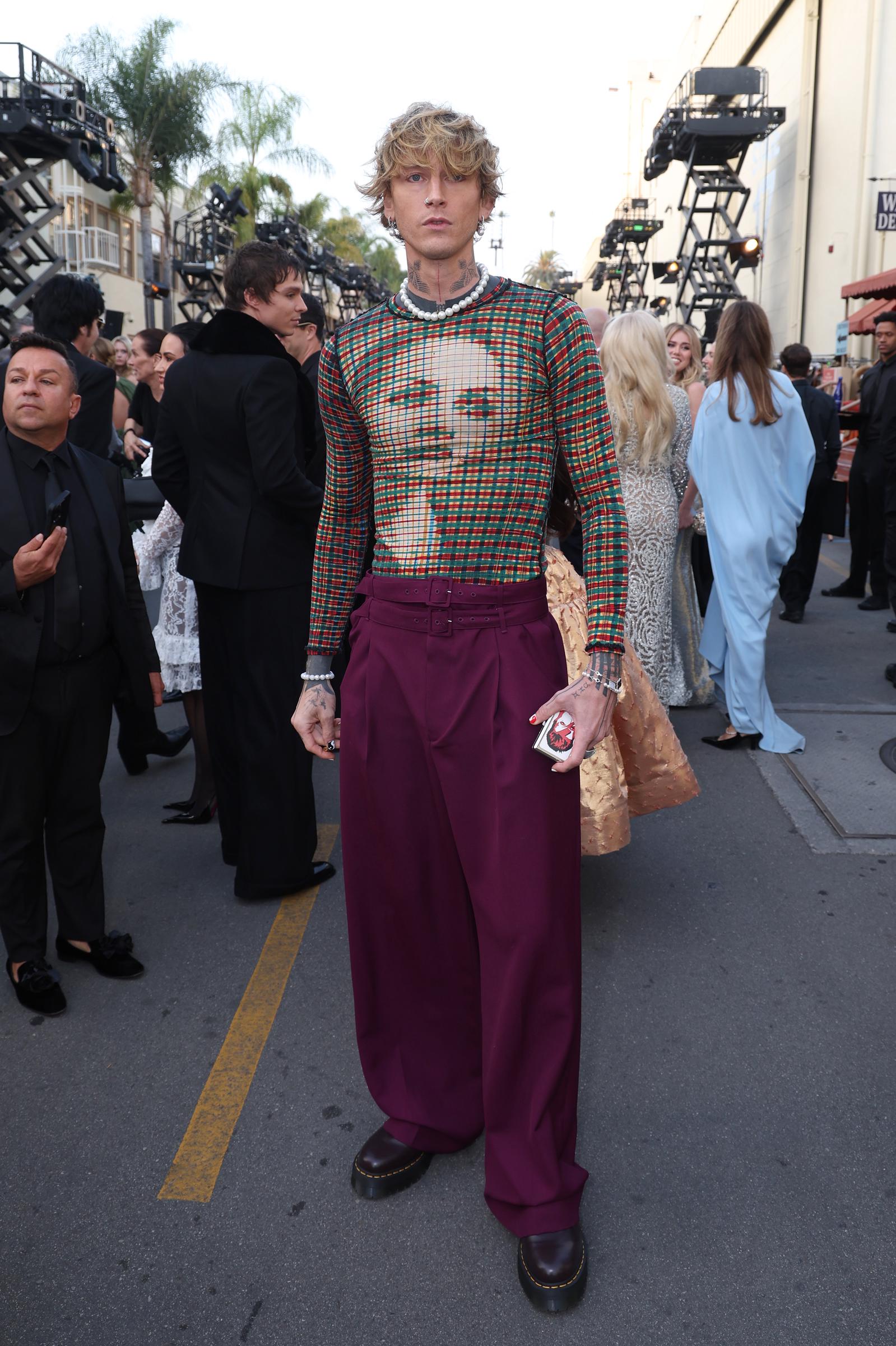 El músico mgk causa sensación con su atrevido look en el evento "Vogue World: Hollywood 2025", celebrado en los estudios Paramount de Los Ángeles el 26 de octubre de 2025. El artista destaca en la alfombra al aire libre con un suéter de manga larga a cuadros, transparente y con estampado gráfico, que combina con un collar de perlas, unos pantalones anchos de color morado ciruela de cintura alta con cinturón y unos zapatos oscuros con suela gruesa. | Fuente: Getty Images