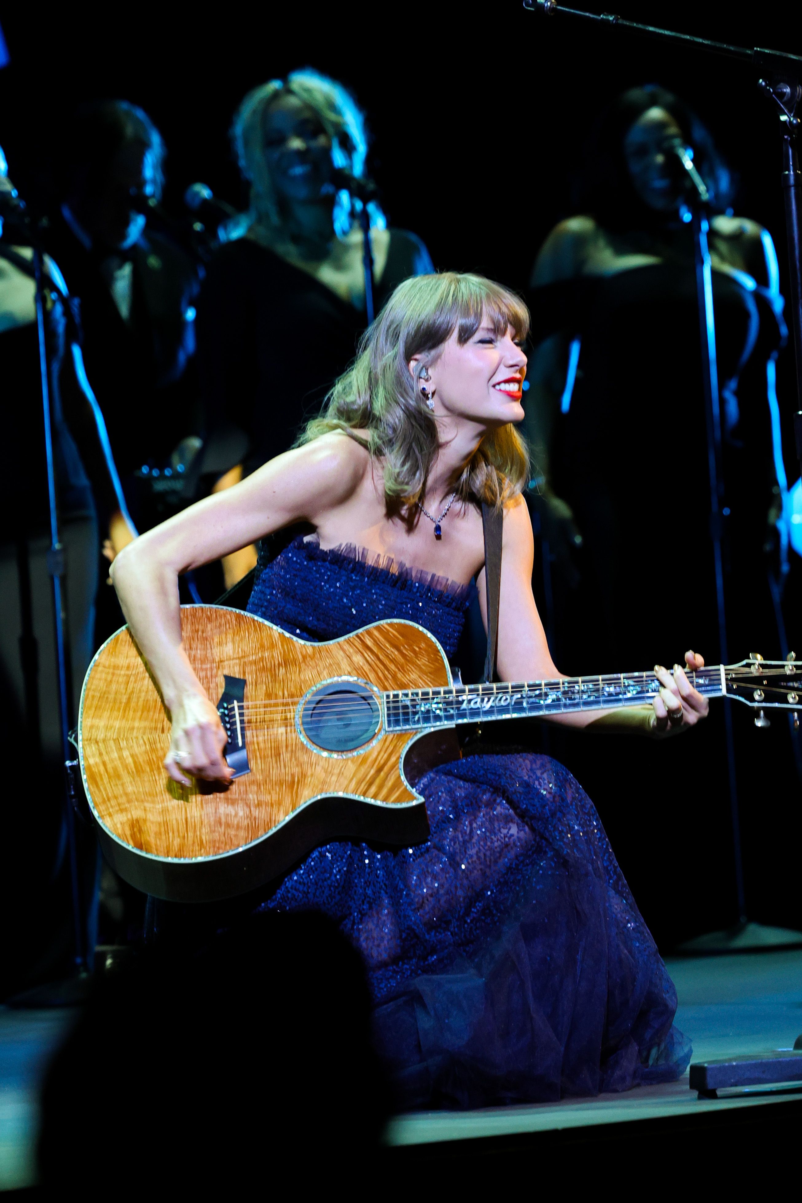 Taylor Swift ofrece un concierto acústico con un vestido azul medianoche sin tirantes, que brilla con lentejuelas por toda la prenda y un delicado ribete de tul en el escote. Sentada con su característica guitarra acústica hecha a medida, la cantante combina este look brillante de escenario con un delicado collar con colgante de zafiro, pendientes de diamantes en forma de gota, su clásico labial rojo y unas suaves ondas de color rubio miel.