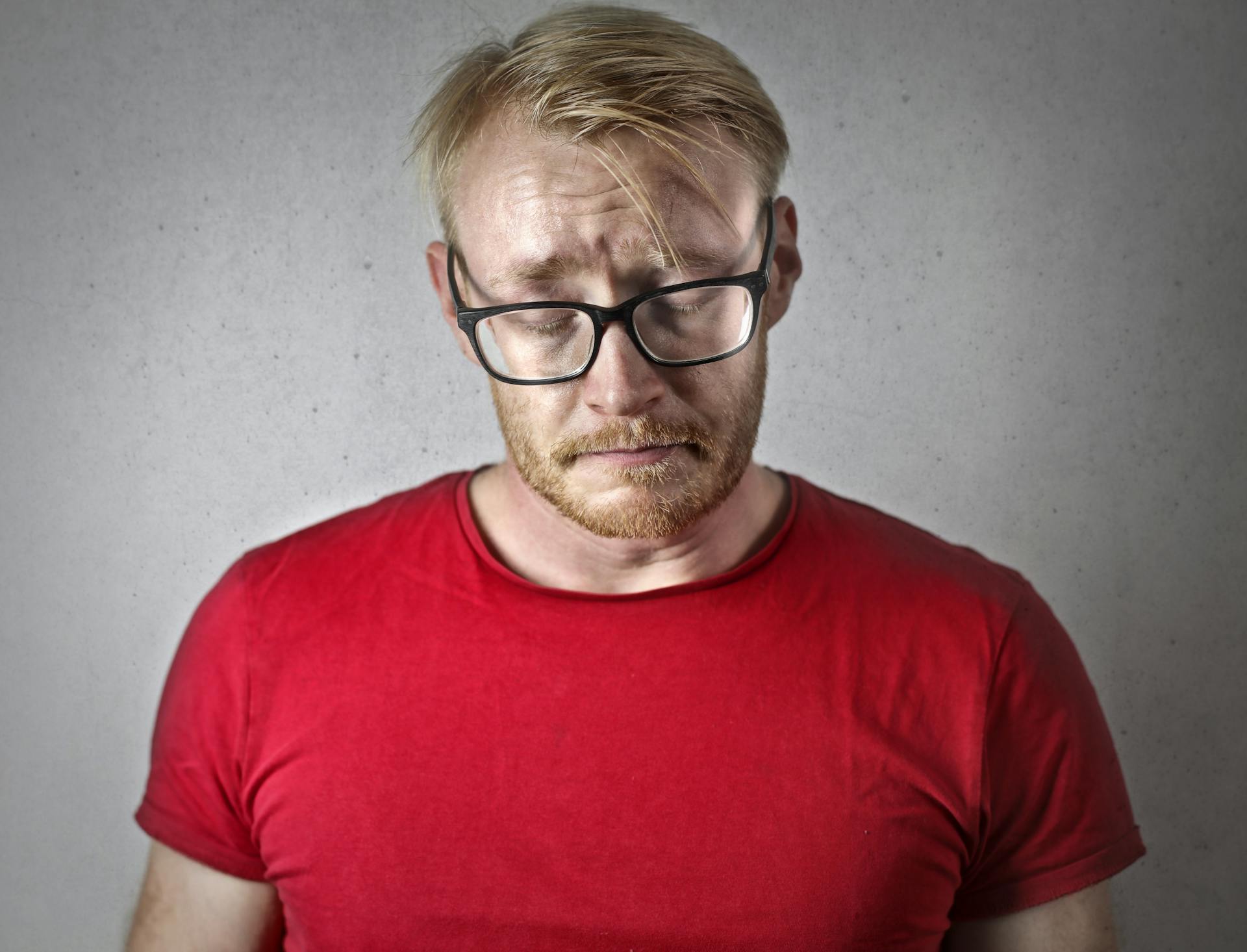 Hombre triste con camiseta roja y gafas de montura negra cerrando los ojos | Fuente: Pexels