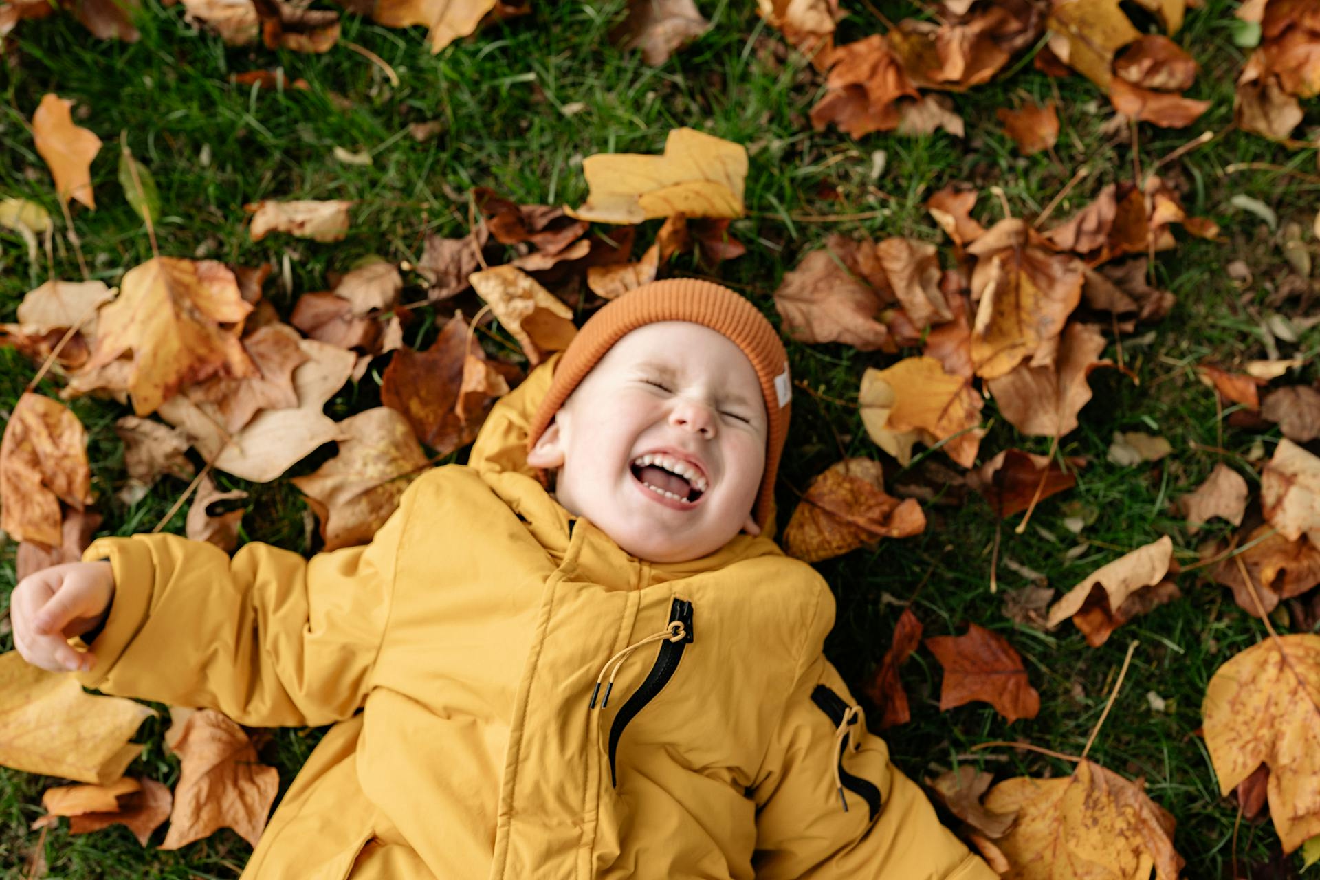 Un niño tumbado en la hierba cubierto de hojas otoñales y riendo | Fuente: Pexels