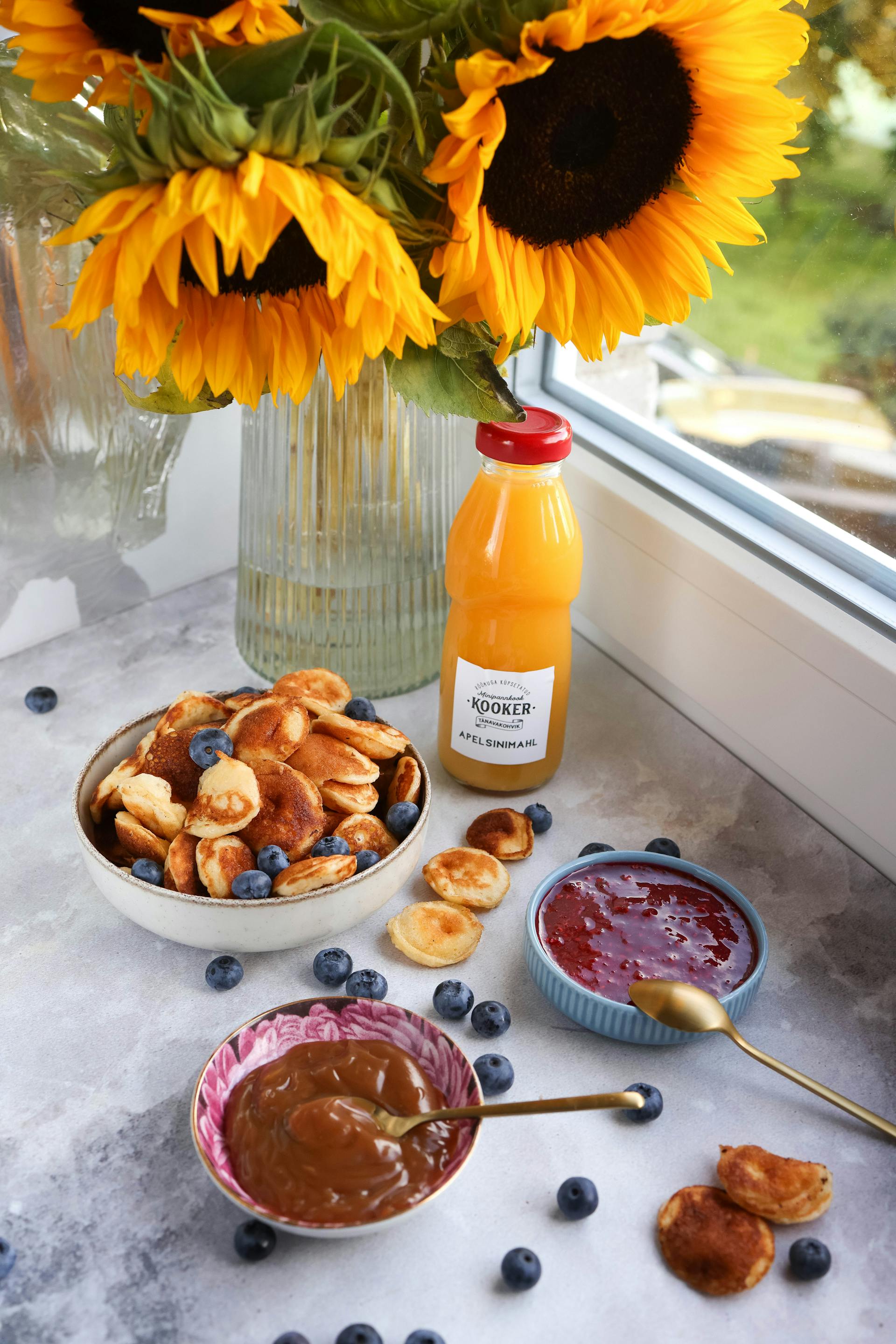 Tortitas con arándanos y zumo de frutas junto a un jarrón de girasoles al lado de la ventana | Fuente: Pexels