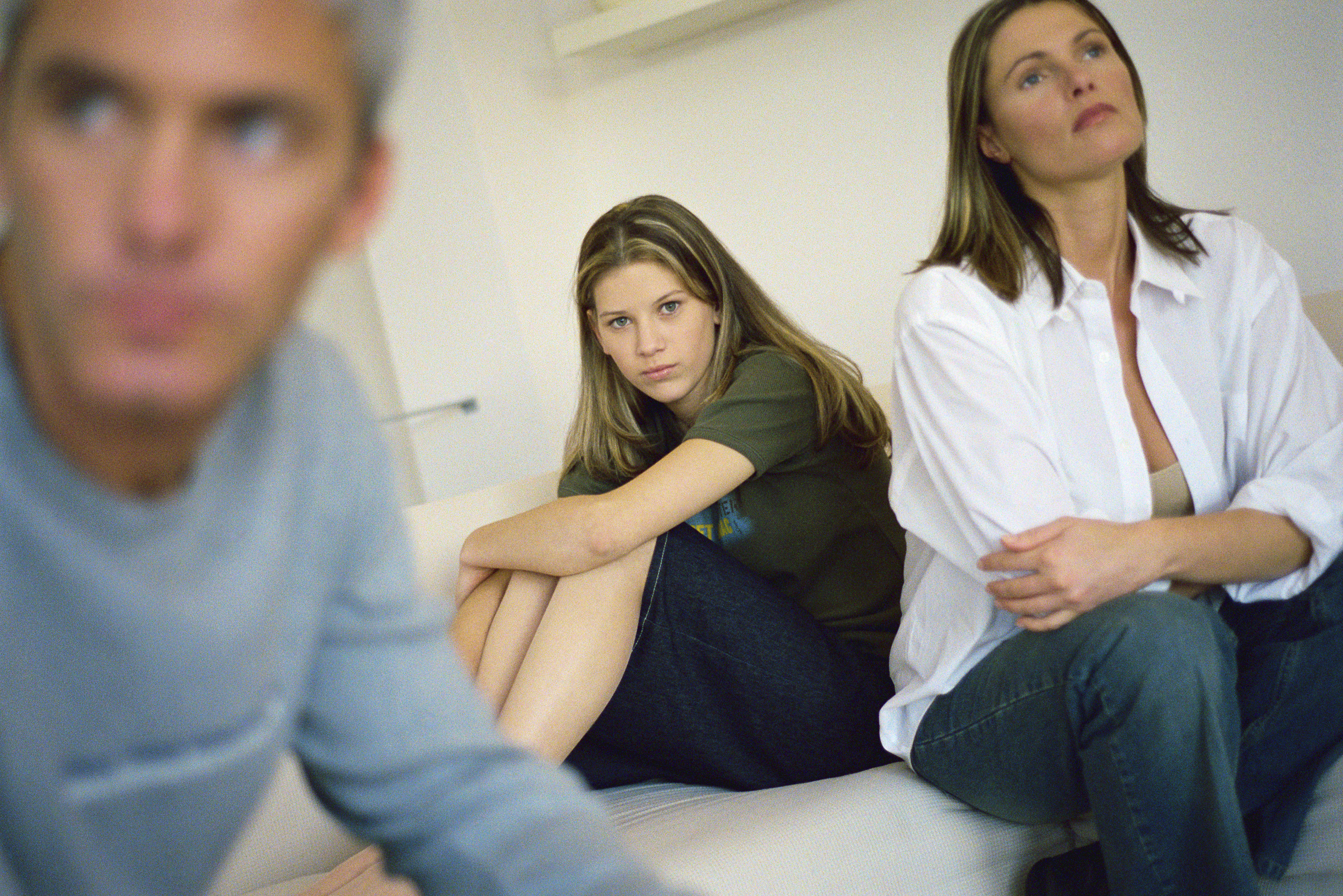 Adolescente sentada en la cama abrazándose las rodillas, los padres sentados cerca mirando hacia otro lado con aire de frustración | Foto: Getty Images