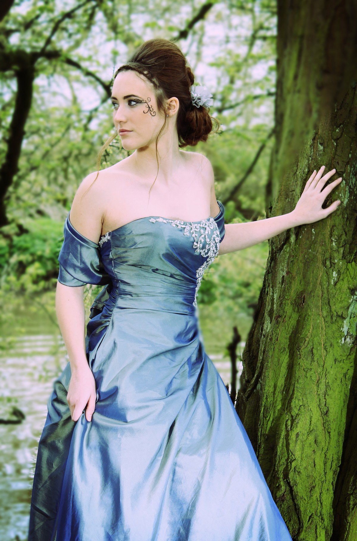 Mujer luciendo un vestido azul y un peinado alto, parada junto a un árbol frente al río. | Imagen: PxHere